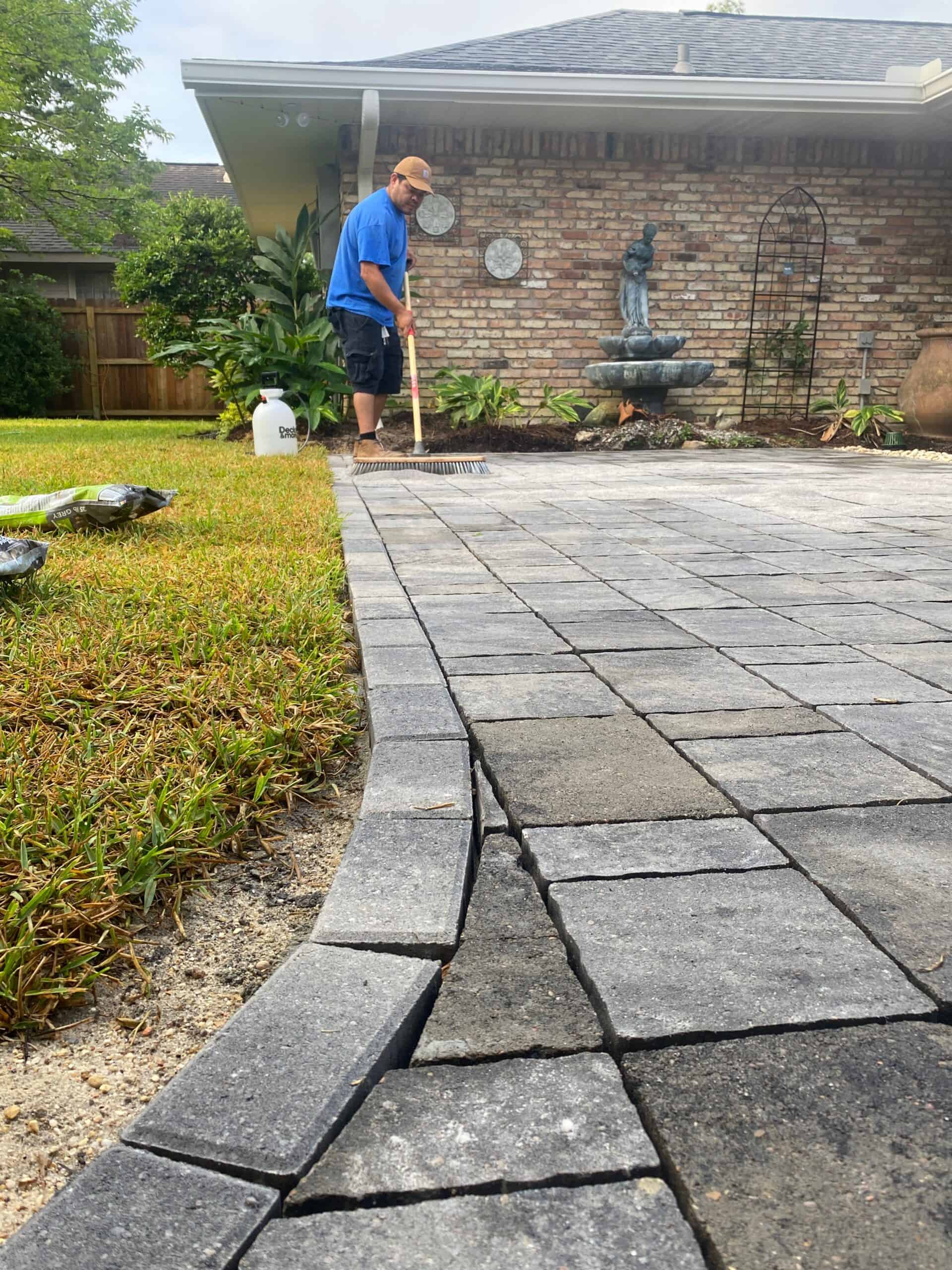 a professional landscaper in a blue shirt using a shop broom to sweet a paver patio in the spring