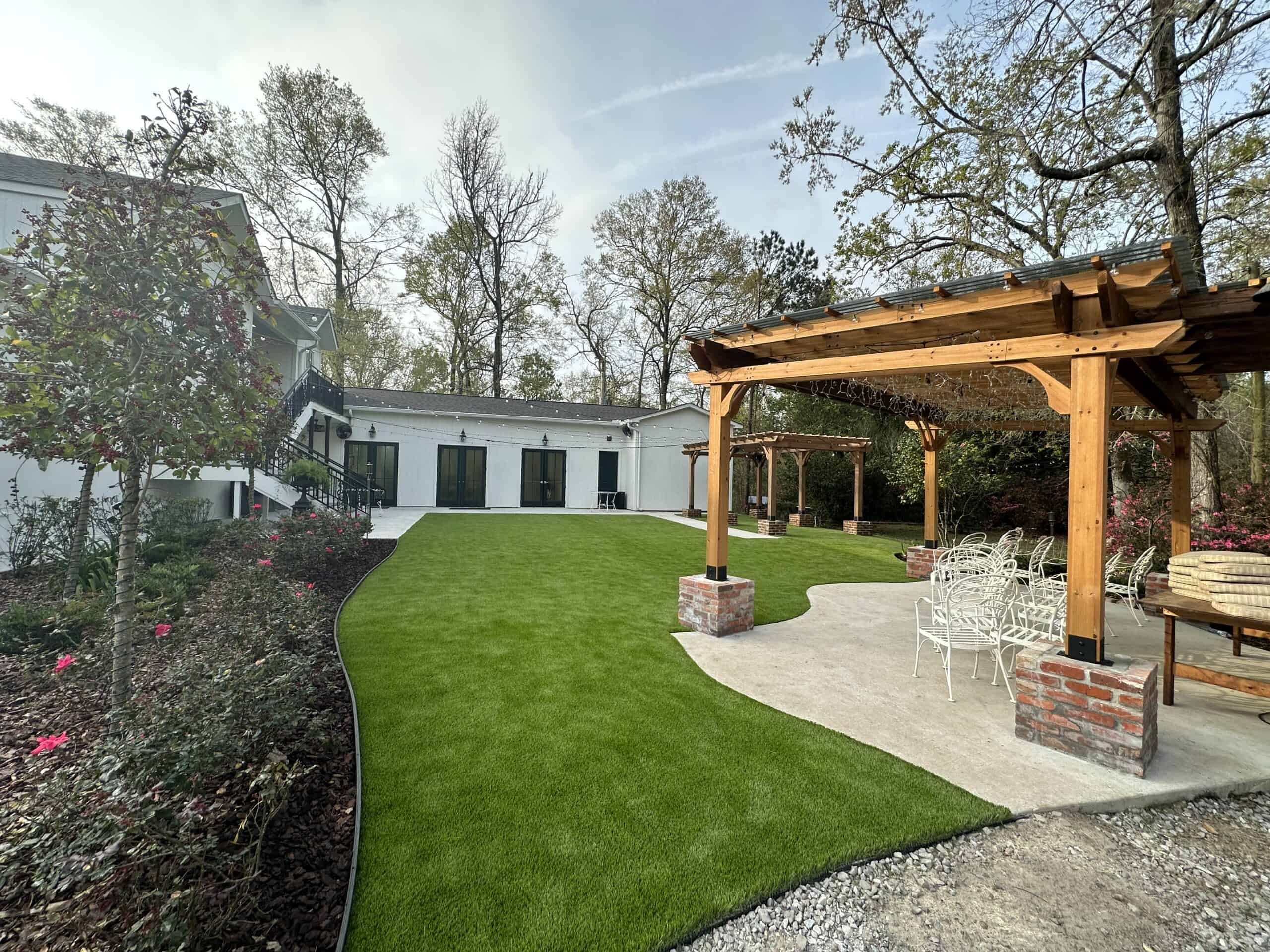 a backyard with artificial turf around pergolas and a mulch bed with flowers and shrubs
