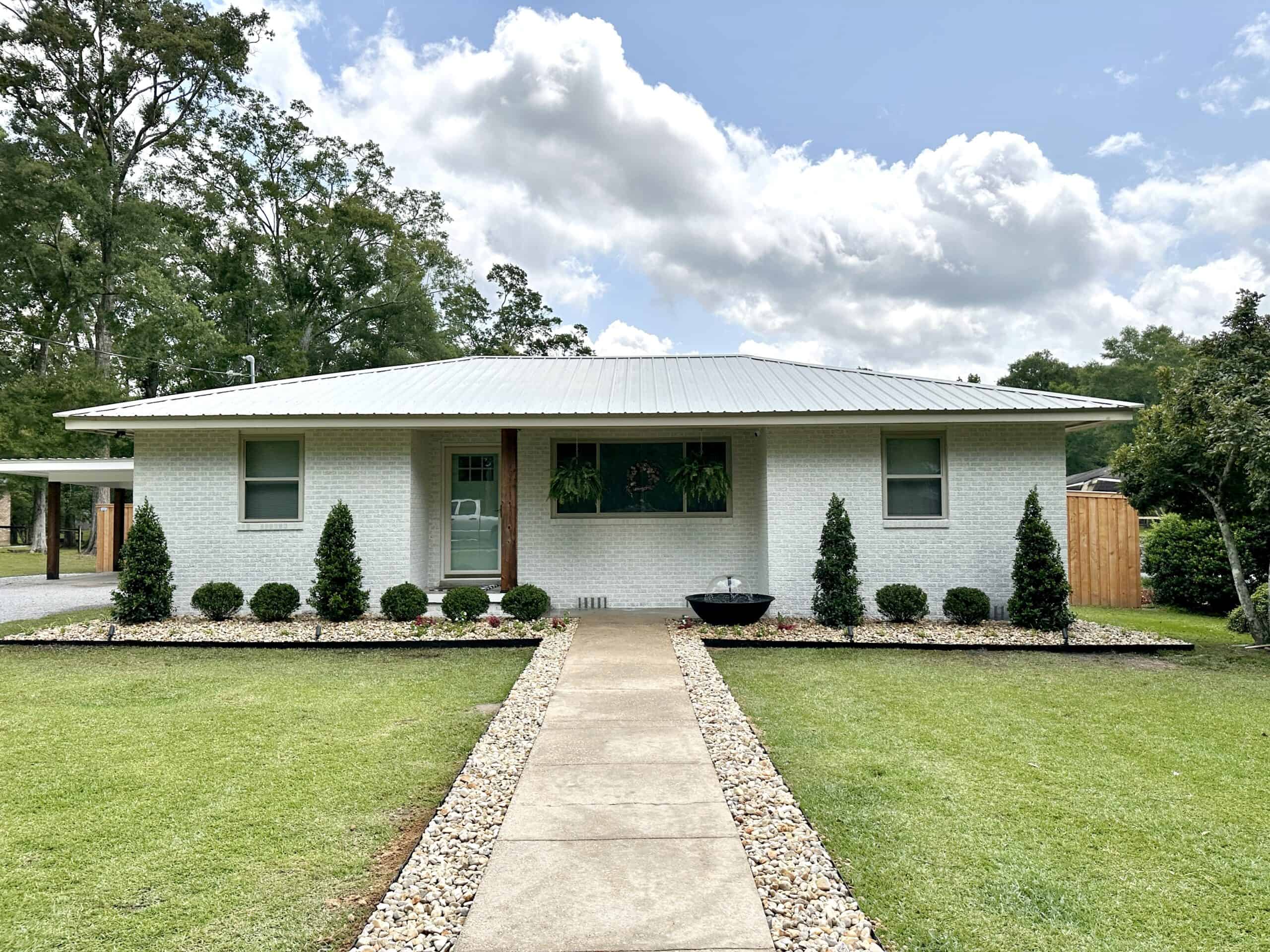 a white brick house with a rock-lined sidewalk leading to a porch surrounded by pruned shrubs